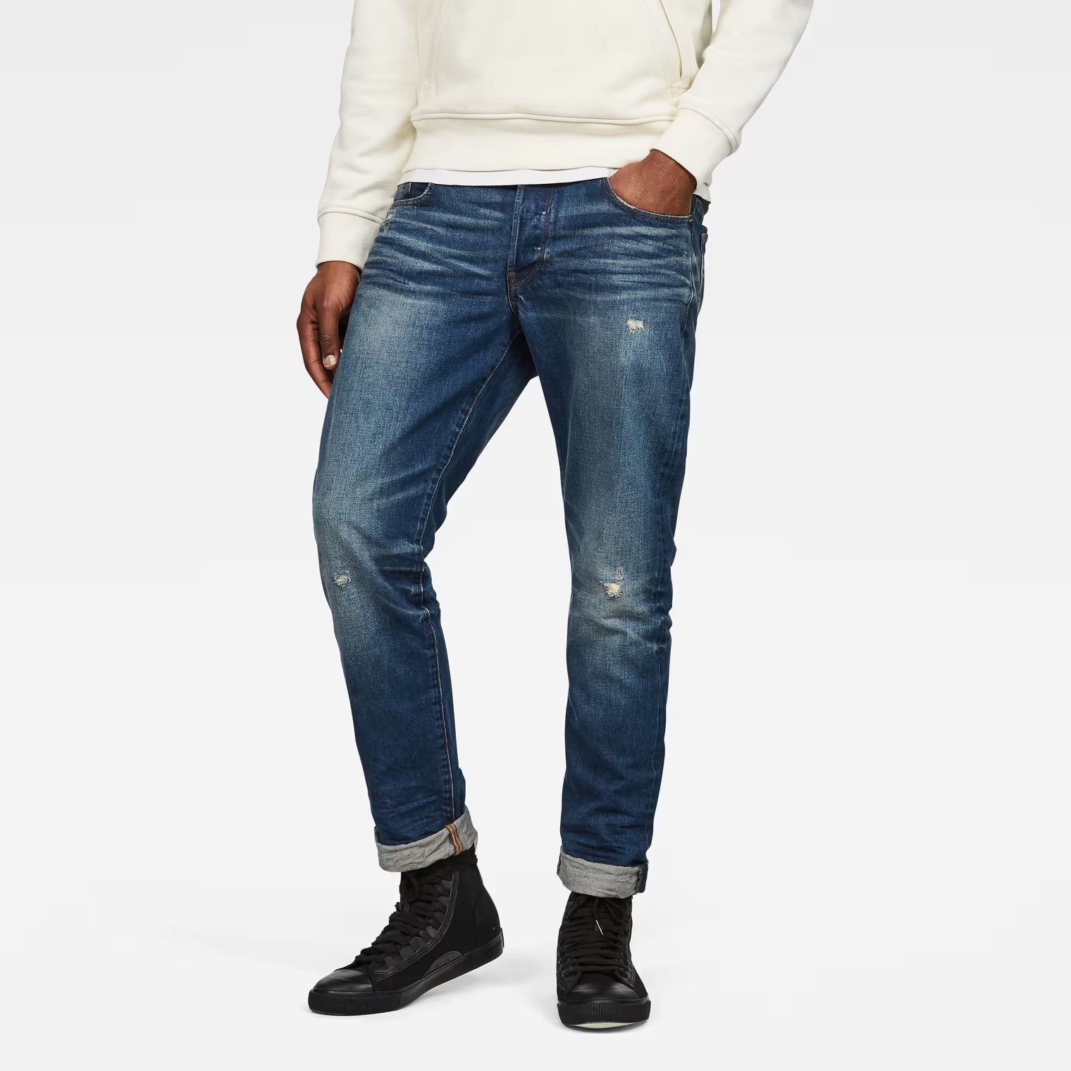 Lifestyle shot of dark blue tapered jeans with distressed details on a white background.