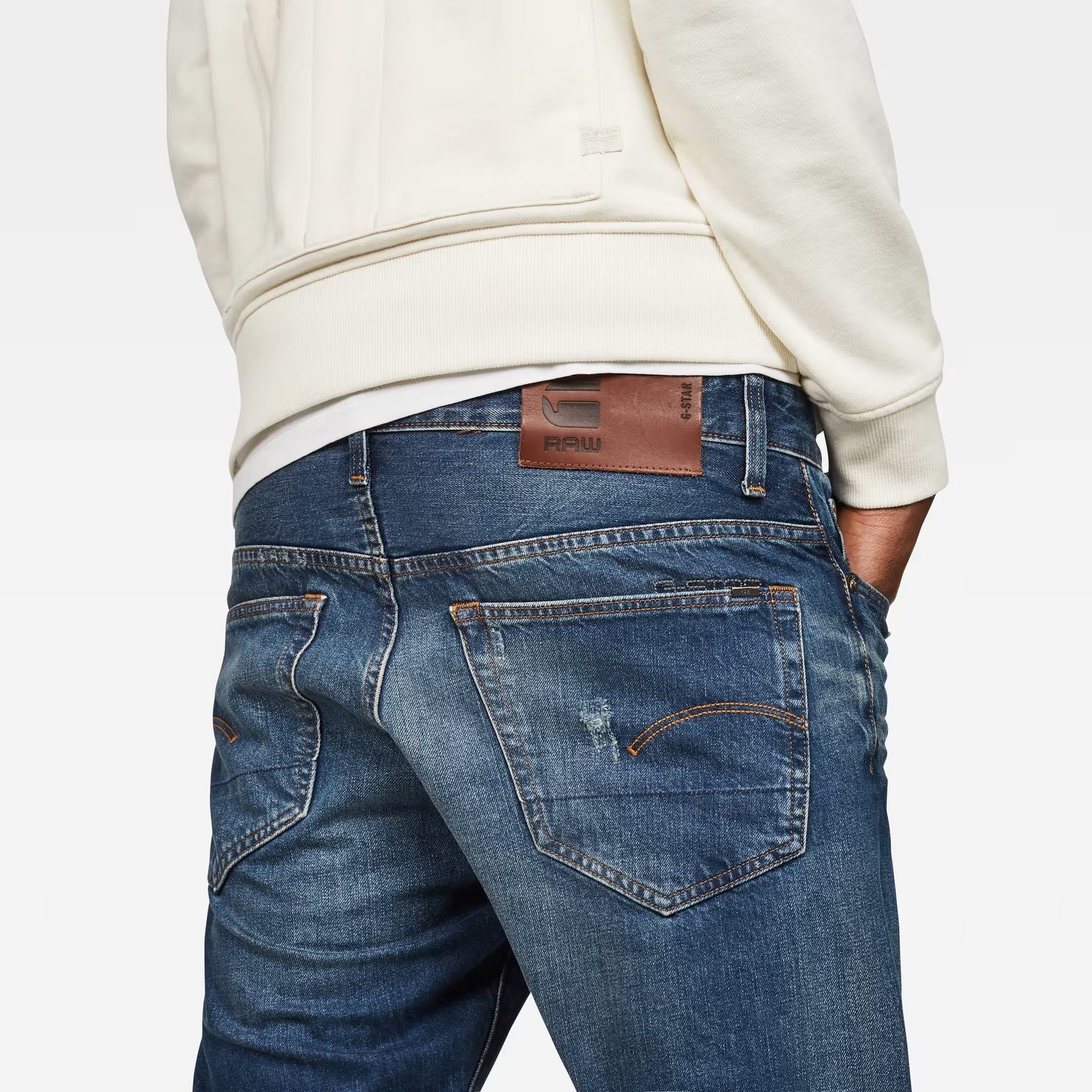 Close-up detail of dark blue tapered jeans showing back pocket and leather patch on a white background.